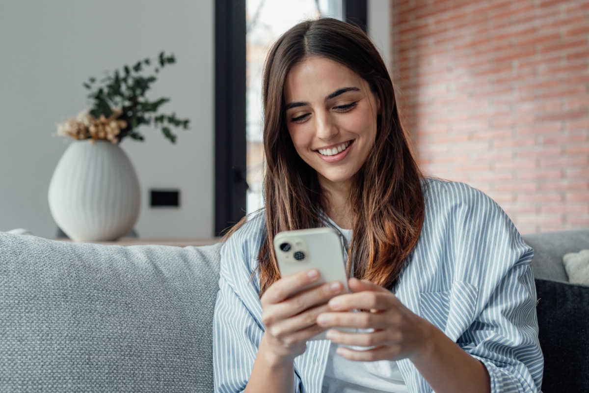 Smiling young adult woman using her smartphone while sitting on the couch.