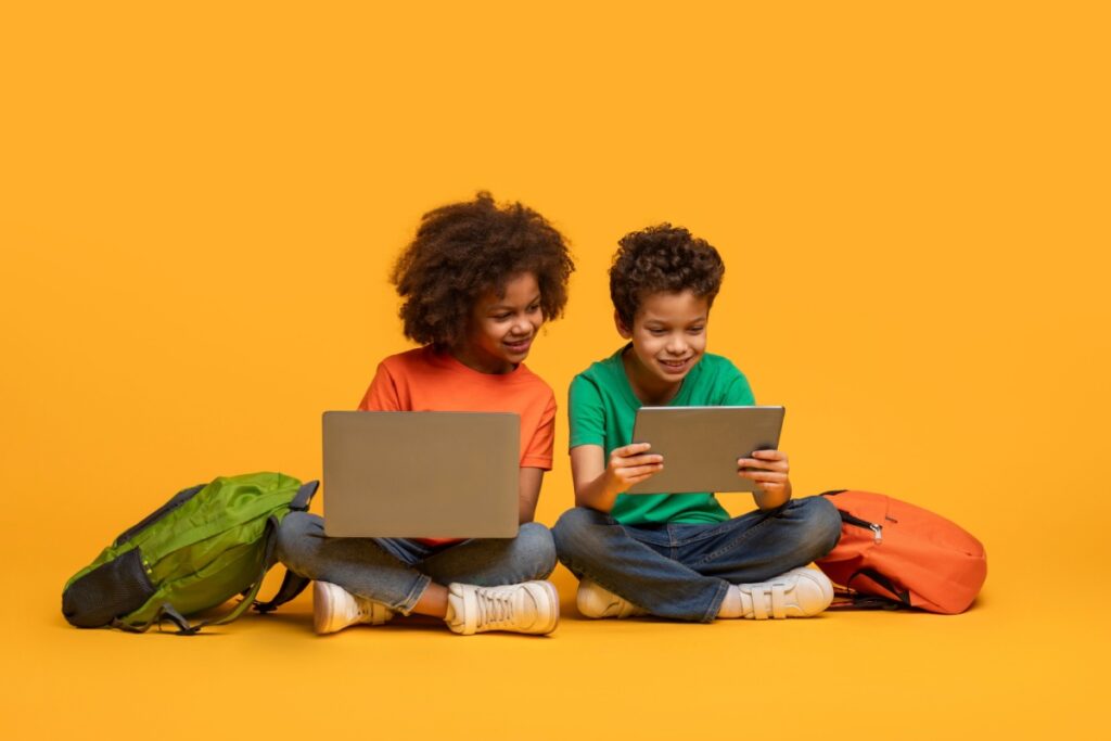 Two ids sitting next to each other using a laptop and tablet.
