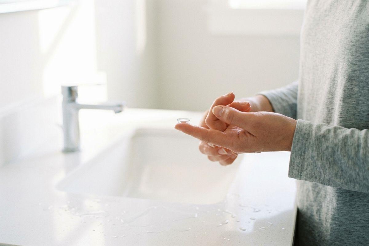 Close-up of a person's hands holding a contact lens on a fingertip above a white bathroom sink with water droplets on the counter.