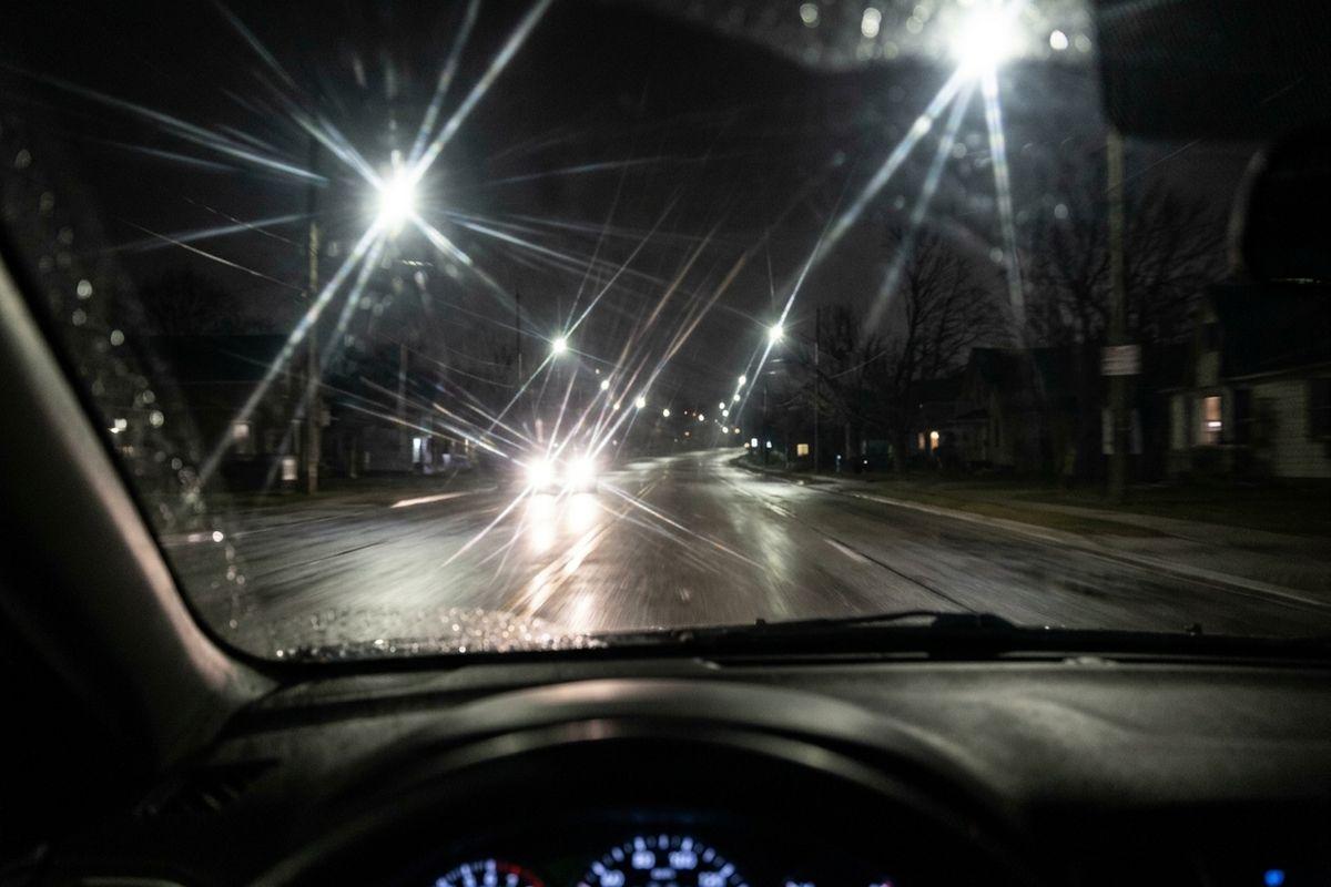View through a car windshield at night, oncoming headlights and streetlights forming starburst glare on a wet road, dashboard visible below